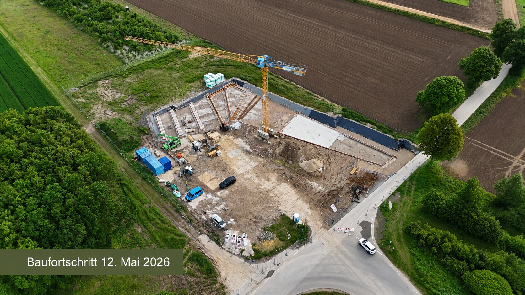 Text: Baufortschritt 12. Mai 2026 - Luftaufnahme des Baufortschritts am Dokumentationszentrum: Auf der Baustelle sind erste Fundamentflächen, Schalungen und ein Baukran zu sehen.