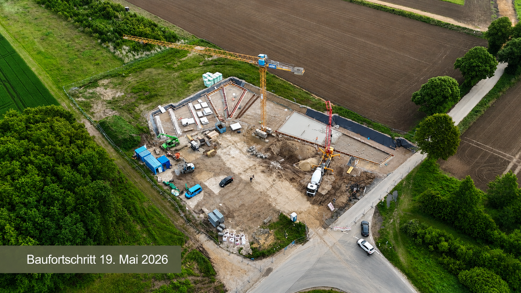Text: Baufortschritt 19. Mai 2026 - Luftaufnahme einer Baustelle für das Dokumentationszentrum. Zu sehen sind Fundamentarbeiten mit teilweise betonierten Bodenplatten, ein Baukran, Betonpumpe und Baufahrzeuge.