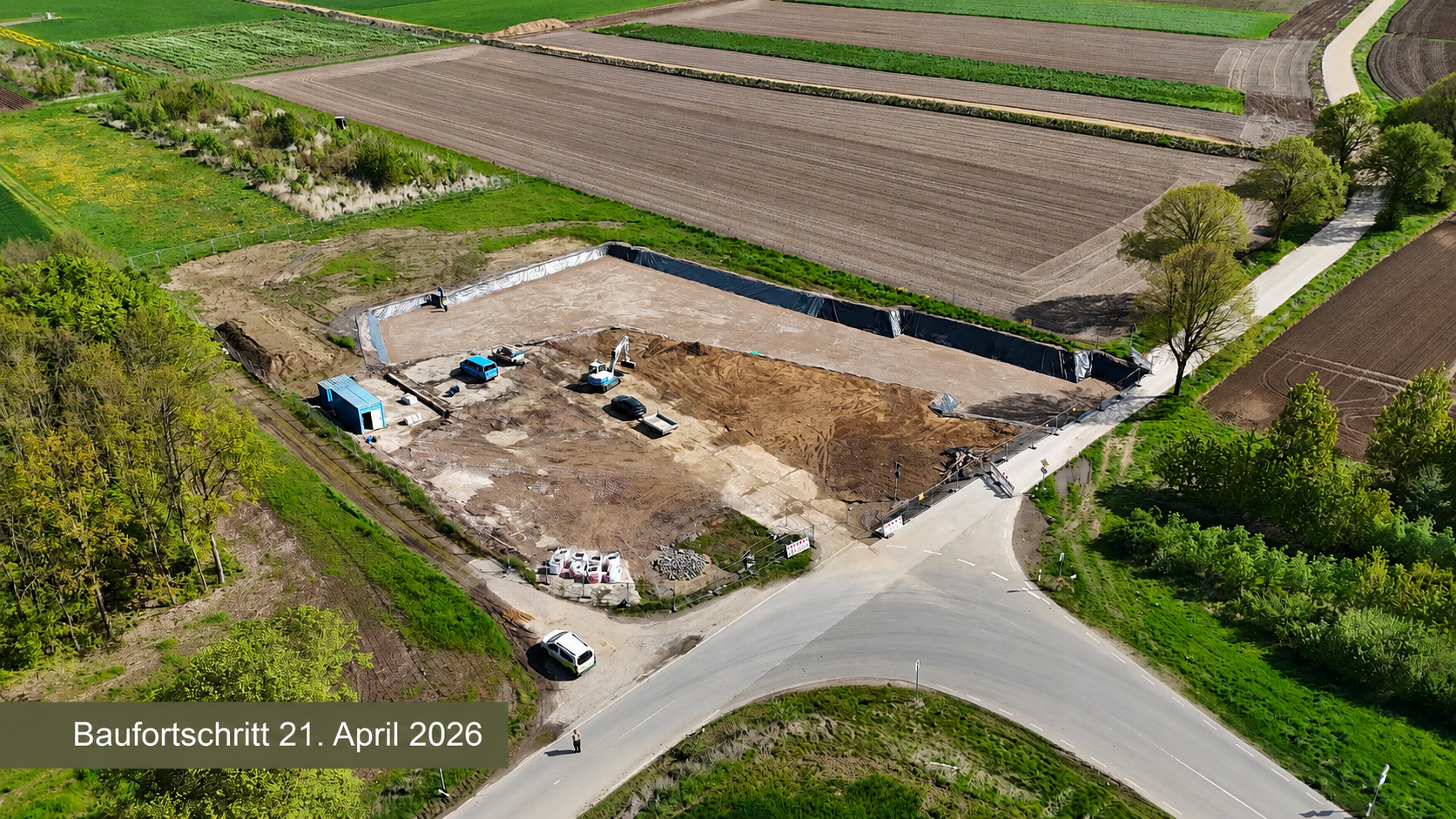 Text: Baufortschritt 21. April 2026 - Luftaufnahme des Baufortschritts am Dokumentationszentrum: Baustellenfläche mit Erdarbeiten, Baggern und einem abgedichteten Baugrubenbereich, umgeben von Feldern und Wirtschaftswegen