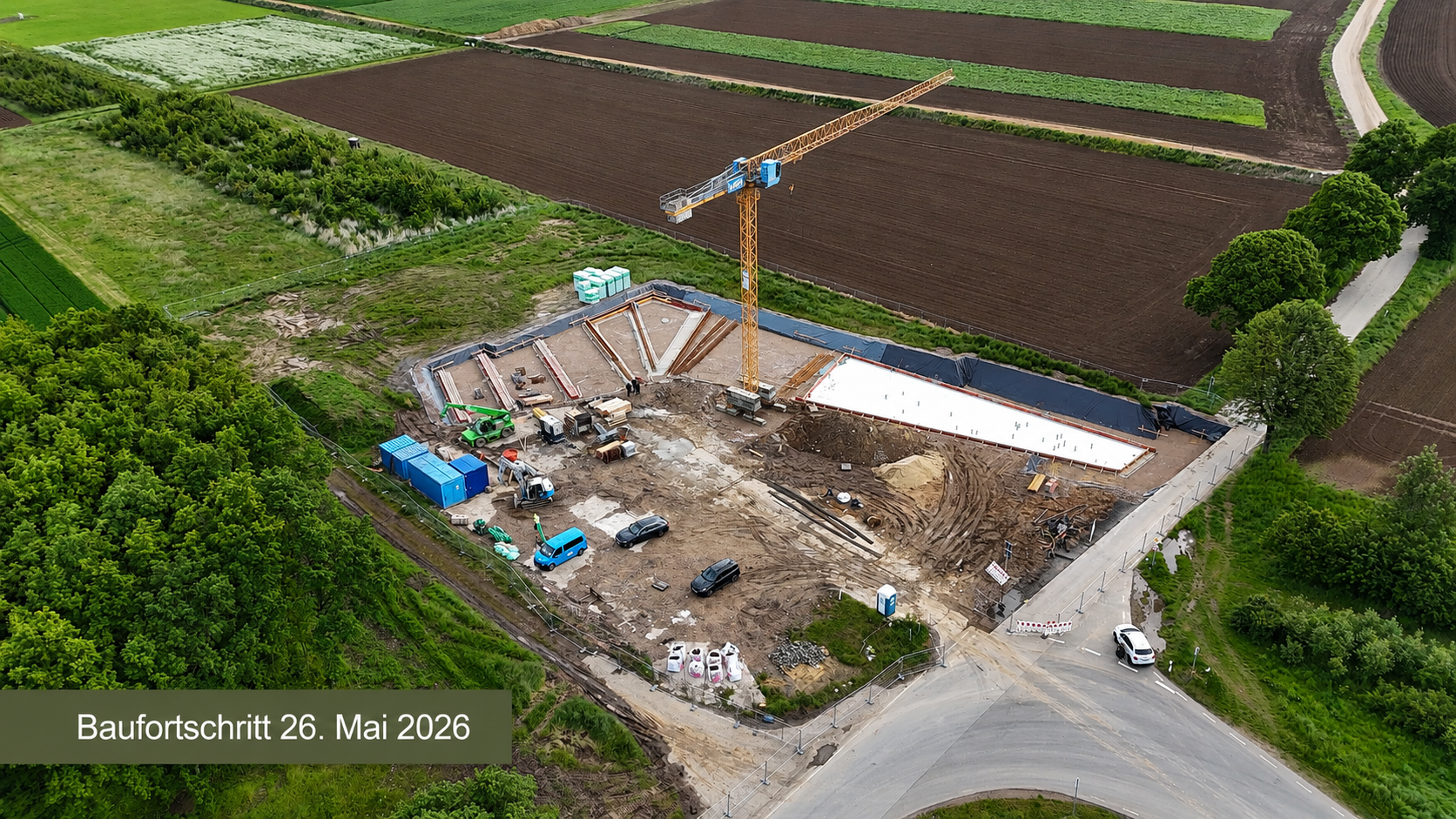 Text: Baufortschritt 26. Mai 2026 - Luftaufnahme der Baustelle des Dokumentationszentrums mit fortgeschrittenen Fundament- und Rohbauarbeiten. Ein Baukran steht über dem teilweise errichteten Gebäude, das von Feldern und Baumreihen umgeben ist. Im Hintergrund ist der Tagebau mit einem Schaufelradbagger zu erkennen.